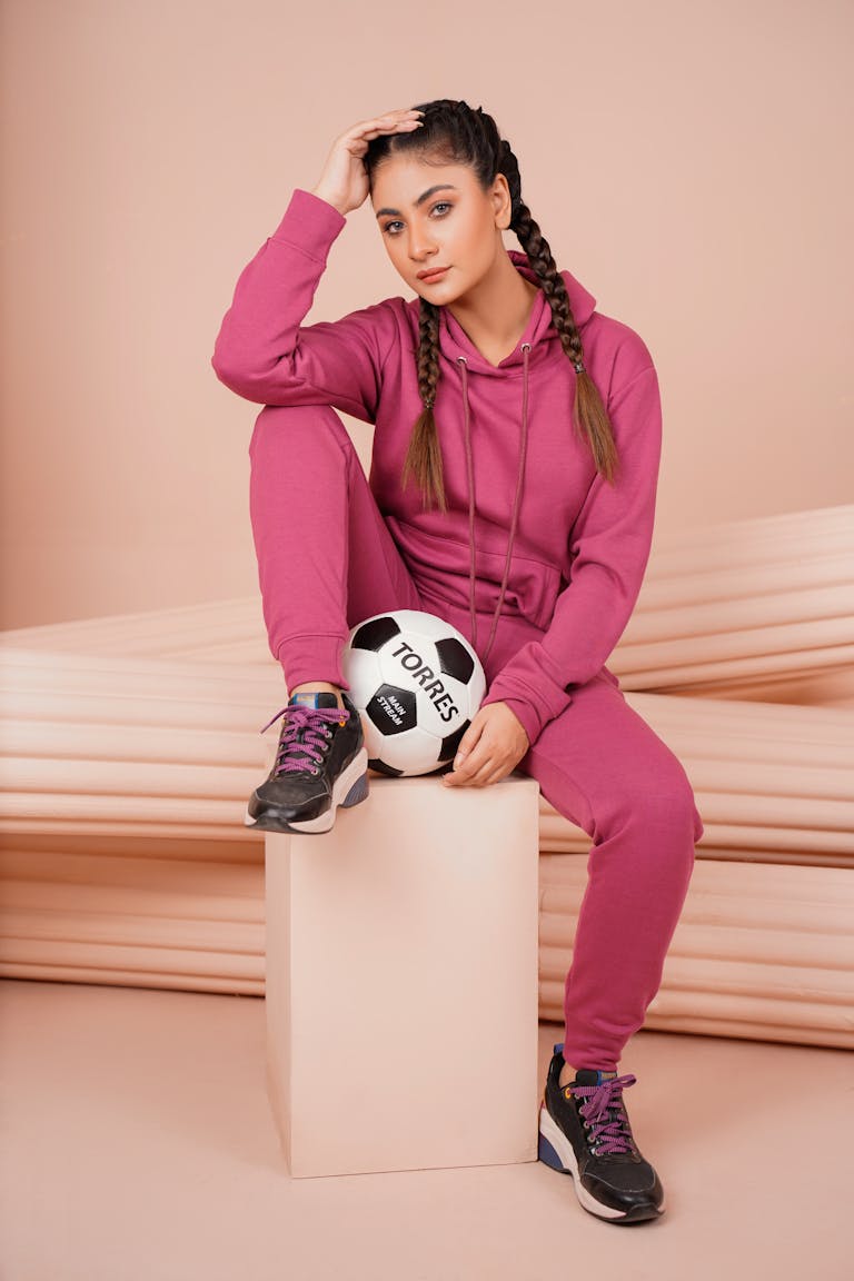 Confident young female in pink athletic wear holding a soccer ball indoors.